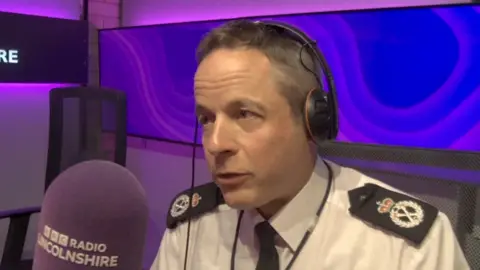 The chief constable speaks into a purple microphone, in a radio studio lit with purple lighting. He wears black headphones and a white shirt with a black tie. He has shirt dark hair with a tint of grey.