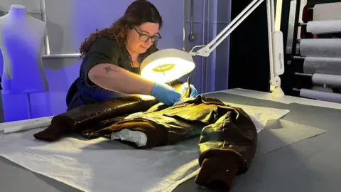 Emma Baugh/BBC A conservator working an airman's flying jacket. She has long curling dark brown hair pulled back from her face and is wearing glasses, a black T-shirt and blue plastic gloves. She is bending over a table where the jacket is lying on its back, with a bright light shining down upon it. It is resting on tissue paper.