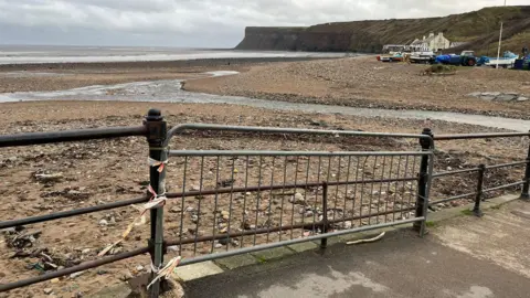 A temporary fence has been attached to the Victorian railing and there are straggly pieces of red and white tape dangling off it. Beyond it is a stony and sandy beach with a channel of shallow water snaking towards the sea. Bots, buildings and cliffs can be seen in the distance.