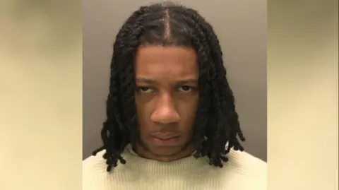 A police custody image of UK rapper Digga D whose name is Rhys Herbert. He has long, braided black hair and dark eyes. He is wearing a cream coloured jumper.