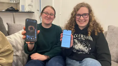 Charley and Bella pictured holding their smartphones up to the camera. Charley is wearing glasses and has brunette hair tied back. Bella has blonde curly hair just past her shoulders.