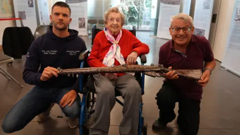 From left to right, Jay Hawkins, Joan Ramsay and John Ramsay are smiling for a picture with the commemorative rifle at the museum.