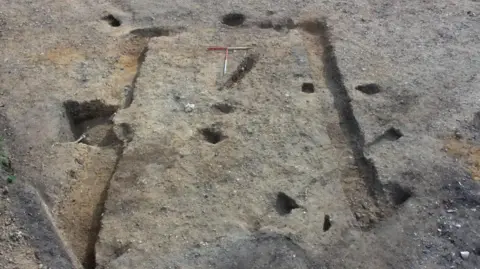 Cotswold Archaeology An excavation of a possible building showing exposed earth and trenches and post holes excavated by archaeologists. The building appears to be rectangular. 