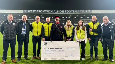 A line-up of nine people with a large cheque on the side of a football pitch. There are seven men and two women. Five of them are wearing lime green tabards. The man in the centre is wearing military uniform.