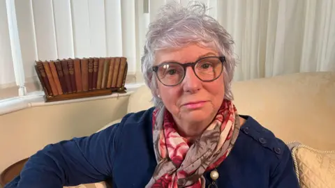 Gill E Andrews sits on a sofa in her home. She is staring at the camera and has short grey hair and thick-rimmed glasses. Ms Andrews is wearing a navy blue jumper and a pink floral scarf. A selection of books is visible in the background.