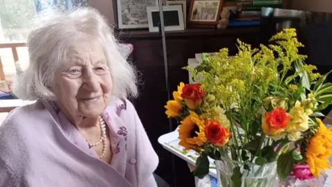 Anne, wearing a lavender cardigan and shirt with pearls, smiles sitting next to a bouquet of flowers, which include sunflowers and roses, in a vase. 