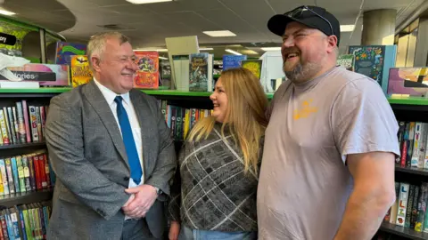 Amy and Ben Grainger stand smiling in a library, with book cases visible behind them. He's wearing a light brown tee-shirt and her a plaid top. They are standing next to Councillor Nick Lakin in a grey suit and blue tie