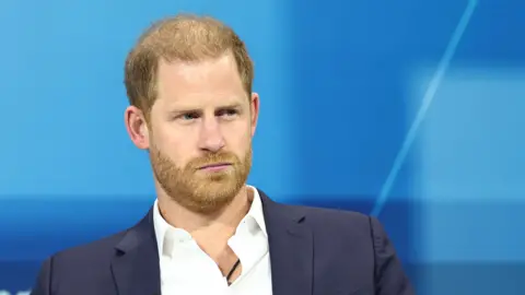 Getty Images Prince Harry wearing a blue suit jacket and white shirt with no tie. There's a blue background behind him and he's looking off into the distance.