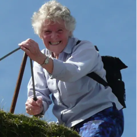 Caitriona MacMillan aunt of Jacqueline Banks- Smiling woman climbing outdoors, holding onto a metal handrail, wearing a light shirt and backpack against a clear blue sky.