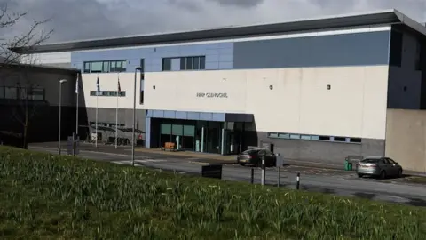 A view of the entrance at HMP Glenochil - the building is blue, white and grey and has two cars parked outside. There is a stretch of grass in the foreground.