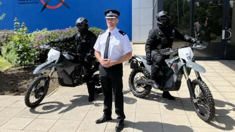 Supt Paul Richardson, wearing a uniform of a white short-sleeved shirt with police patches, black tie and trousers and a police officer's cap, standing between two riders on the new, lightweight electric motorbikes which have a similar shape and design to motocross bikes. The riders themselves wear all black, including blacked out helmets.