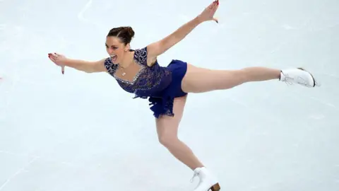 Jenna McCorkell has her brown hair up bun. Her arms are outstretched and she is skating on one skate. She is wearing a dark blue ice-dancing dress which is covered in sequins. 