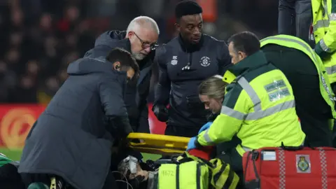 Reuters Luton Town's Tom Lockyer receives medical attention on the field after collapsing during a Premier League match. Tom's head can be seen on the floor with an oxygen mask over his mouth. There are two paramedics in high-vis tending to him and he is surrounded by medical equipment and a stretcher. There are also three members of Luton Town's medical team around him and they are all wearing dark blue coats