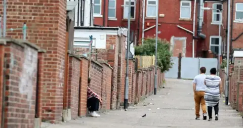 Backstreets and alleys in the Eastwood area of Rotherham, with a man and a woman walking, and a man sitting. A bird and some litter is in the street with many red brick buildings.