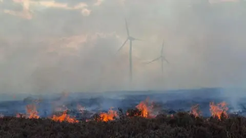 West Yorkshire Fire and Rescue Service Bright orange flames burning on the moorland amid rising smoke; two wind turbines can be seen in the background