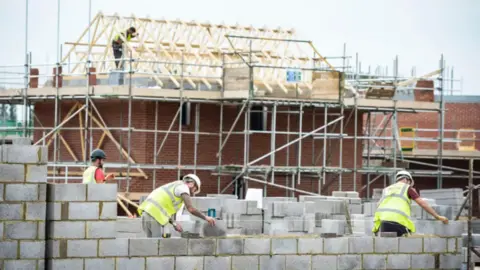 Builders on a housing site. In the foreground three builders cement bricks together. In the background a builder stands on top of a mostly built red brick house, with the exposed wooden roof beams also in place. 