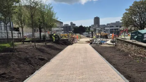 Granite paving along Armada Way. Soil lines either side of the pathway. A worker, dressed in a hi-vis tabard, is stood on the soil. Trees have been planted. Machinery can be seen on the site. Plymouth's Civic Centre is in the distance.
