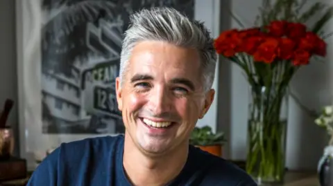 Donal Skehan has salt and pepper hair, parted to the side and raised from his forehead. He is wearing a navy t-shirt and a vase of red flowers is behind where he is sitting