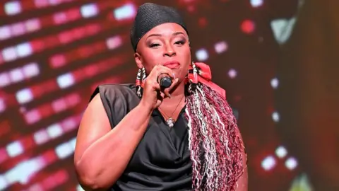 Getty Images Jaguar Wright pictured performing in Atlanta in September 2024. The background is flashing red lights and she has a black head piece, black sleeveless top and large hoop earrings.