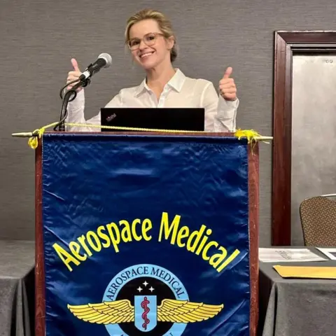 Christina Mackaill Christina Mackaill standing at a podium behind a flag reading AEROSPACE MEDICAL ASSOCIATION. She is holding her thumbs up wearing glasses and a white blouse.