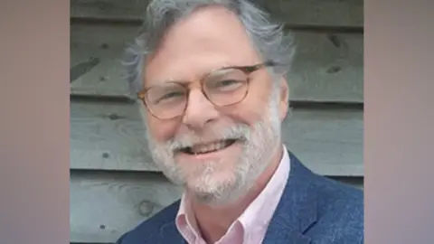 Ashdown Forest Foundation Robin St Clair Jones is pictured wearing glasses and a jacket, standing in front of a wooden fence or wall.