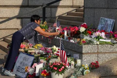 Getty Images A display of flags and flowers fills the image as a woman leans over to lay a flower in the middle of the arrangement