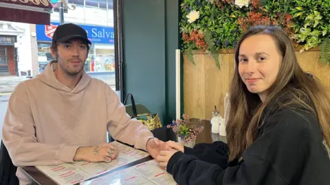 A young couple sit at a table in a cafe holding hands