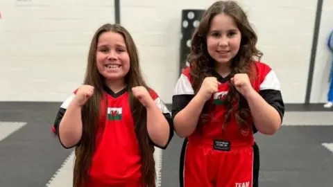 Debra Davies Lili-Rose and Annarose face the camera in their Team Wales tracksuits, smiling and with arms raised 