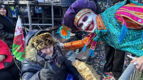 Photograph of Avril Davidge, sat outside in her wheelchair. A Welsh flag can be seen poking out of her wheelchair. She wears a leopard furry hat, a purple scarf and a black puffer coat. She wears small reading glasses and holds her thumb up to the camera. A captain of the Mummers parade leans down to her and smiles. He wears a vibrant blue, purple and orange sequined costume and has a large red joker smile painted on his face. 