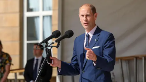 ED CRISPIN/DUKE OF EDINBURGH'S AWARD Prince Edward with short grey hair, wearing a blue jacket and tie, holding his hands forward and speaking into two microphones on a stand. There is a man in a suit standing in the background and the light stone of Buckingham Palace is visible behind.