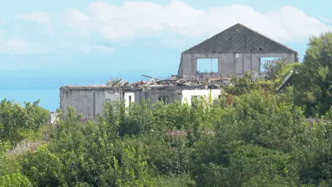 The Idlerocks Hotel, a building that is derelict, with a facade on the far side and two empty windows. The building is surrounded by greenery. Behind it is the sea. 