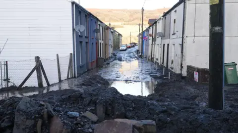 A pile of sludge and mud blocks the entrance to a road which is covered in water. The mountain is visible in the background behind a row of terraced houses.