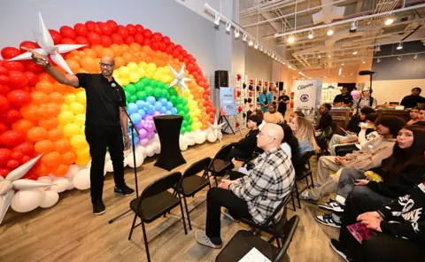 Adam Pantozzi / NBAE via Getty Images Collins gestures while speaking in front of a balloon installation in the colours of the LGBT+ flag while people sit on chairs in front of him listening, inside a convention centre.