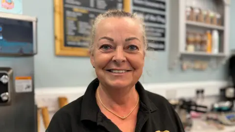 A head and shoulders photograph of Tracy Walters in her sweet shop. She has blonde hair that is pulled back in a ponytail and she is wearing a black T-shirt.
