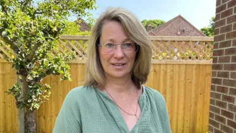 Shaun Whitmore/BBC Christine Cullen, wearing a green blouse and a chain around her neck, stands in a garden. She is wearing glasses and has shoulder-length blonde hair. Behind her is a wooden fence and a tree.