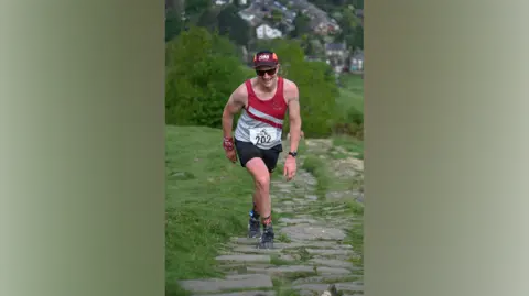 Rob Hasler A man wearing a running vest with a number on it, and a cap and sunglasses, walks up a large stoned hill with grass either side 