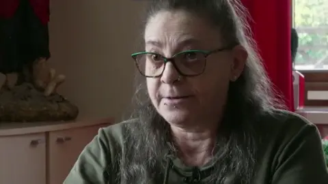 Aisha Iqbal/BBC A middle-aged woman with long brown hair, glasses and wearing a dark green top, sits in a kitchen.