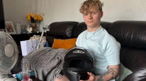 George King/BBC Harry Pyett sitting on a black leather sofa holding his black crash helmet. Next to him is a table with a fan and a glass of drink on it. On the left side of the sofa is a blankets and his crutch. He is looking into the camera.