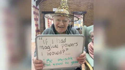 Temple Croft Care Home An elderly man sitting down, smiling and wearing a witches hat holding up a sign that says "if I had magical powers I would?" with the answer "be invisible, that would be good".