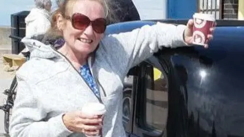 Merseyside Police A woman in her late 60s smiles at the camera while leaning against a black cab. She is holding two hot drinks in disposable paper cups in each hand, has light ginger hair tied back and is wearing a grey zip-up hoody and sunglasses.