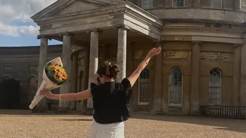 Contributed A woman has her back turned to the camera toward a large stately home. She has her arms raised in celebration. In one hand she holds a bunch of sunflowers. She has dark hair that has been tied back and she wears a black blouse with a white skirt. 
