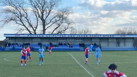 Juliette Parkin A football stand has the words "Matthew Grimstone and Jacob Schilt stand" at the top. There are people seated, watching a match, and footballers play in front of them. A large tree rises above the stand. the sky is blue with some white clouds.