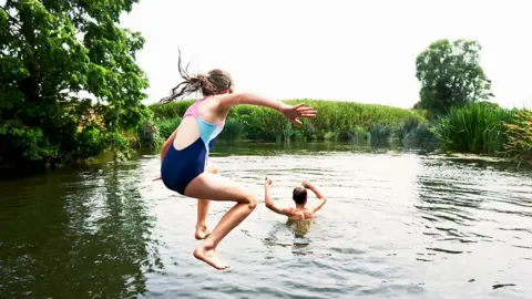 A boy and a girl jumping into a lake surrounded by trees and reeds