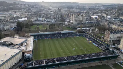 Getty Images The Recreation Ground in Bath seen from above. There is a rugby game going on and the stands are filled with people. Behind the stadium are the hills of Bath.