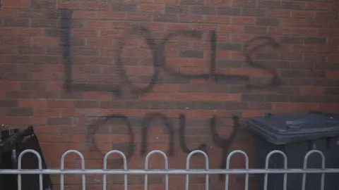 Black spray paint has been used on a red brick wall. It should read "Locals only", however has been mis-spelled to say LOCLS only. A black wheelie bin is also visible behind a low metal fence. 