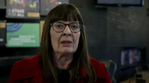 A woman with brown hair to just below the shoulders and strong, defined fringe, is staring into a camera. She is wearing glasses that have a brown rim and is wearing a red jacket over a dark top.