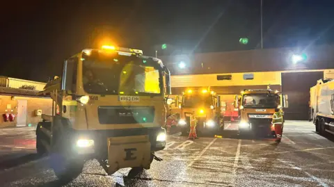 Dorset Council Several yellow gritters with lights on at night.
