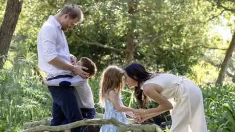 Archie is hugging his father, Prince Harry, the Duke of Sussex, while Meghan is bent to eye-level and holding hands with Lilibet, as they all stand on what appears to be carved mini wooden bridge over a brook among trees.