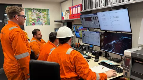A group of men in orange overalls looks at computer screens