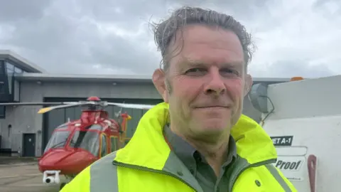 A middle aged man with grey hair that is long on top looking straight at the camera, you can see his head and shoulders and he is not smiling. He is standing outside an aircraft hangar wearing a fluorescent jacket and a grey shirt. In the background you can see part of a white fuel tank and a red and yellow Cornwall Air Ambulance on the ground.